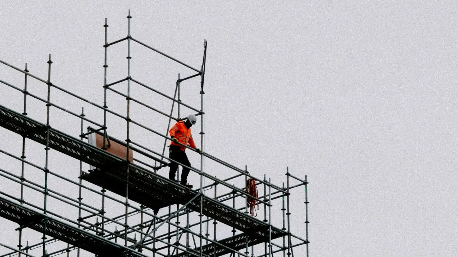 A lone worker high on scaffolding, seen from a distance against a grey sky.